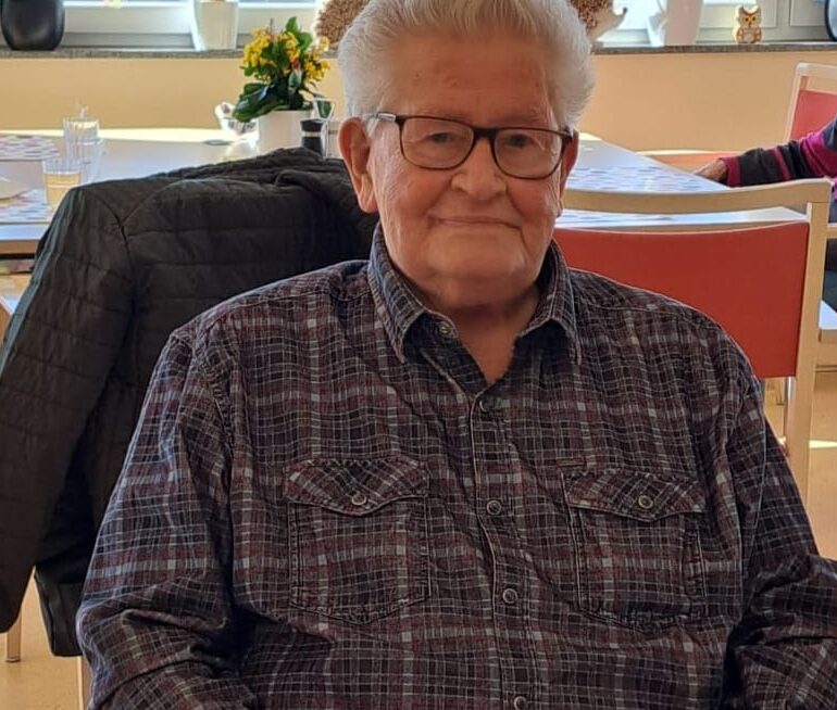 Senior mit Brille sitzt lächelnd im hellen Seniorenzentrum; Tisch mit bunten Punkten, Pflanzen am Fenster.