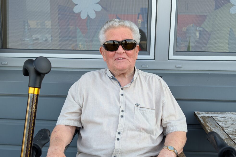 Senior mit Sonnenbrille sitzt entspannt auf Balkon vor Fenster, neben Rollator und Gehstock, Sommertag