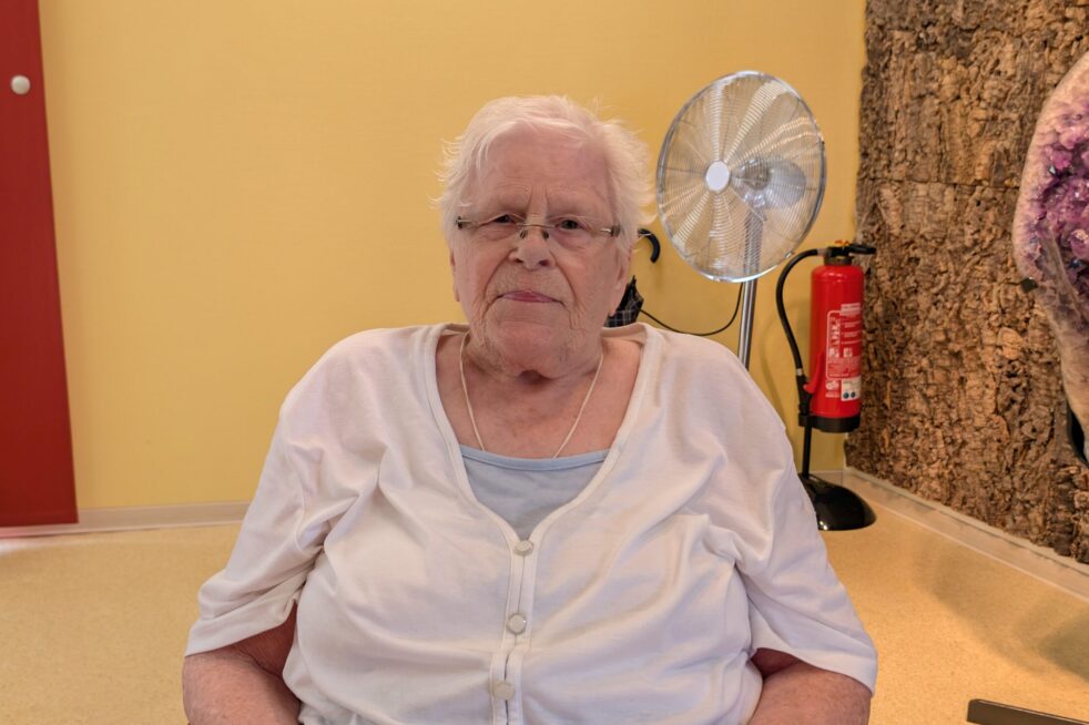 Seniorin sitzt in einem hellen Raum auf Stuhl; gelbe Wand, Ventilator und Feuerlöscher im Hintergrund.