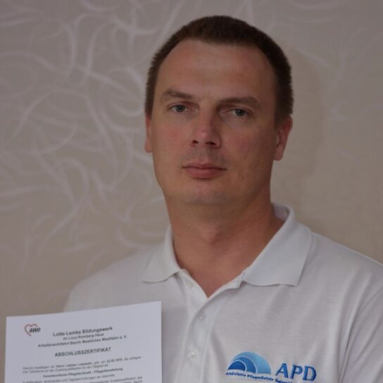 Mann im weißen Poloshirt mit APD-Logo zeigt Abschlusszertifikat vor neutralem Hintergrund.