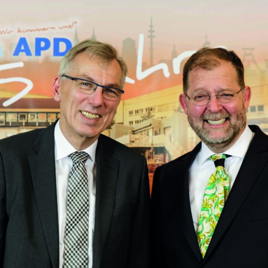 Zwei Männer im Anzug lächeln vor APD-Banner beim 25-Jahre-Jubiläum des Unternehmens.