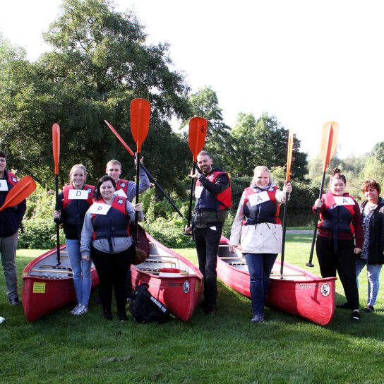 Team beim Kanufahren: Gruppe mit Rettungswesten und Paddeln vor roten Kanus im Grünen beim Teambuilding.