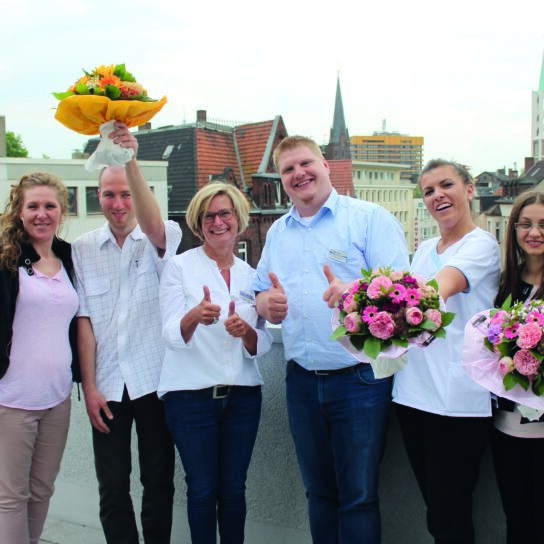Sechs Teammitglieder auf Dachterrasse, fröhlich mit Blumensträußen, Daumen hoch, Stadtpanorama im Hintergrund