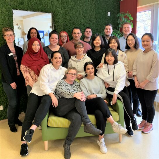 Gruppenfoto eines vielfältigen Mitarbeiterteams, lächelnd im Büro auf grünem Sofa vor Mooswand.