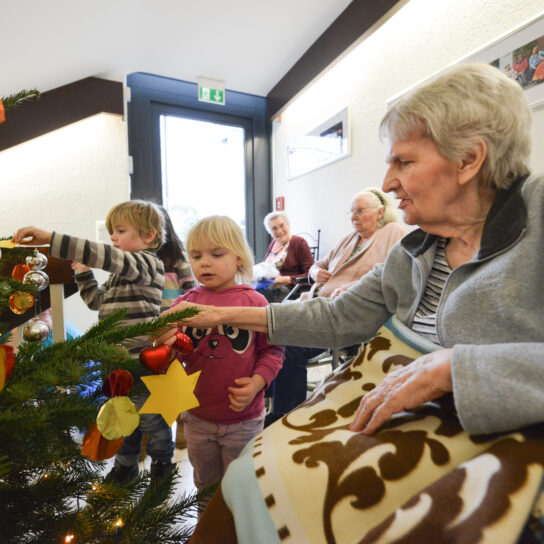 Kinder und Senioren schmücken gemeinsam den Weihnachtsbaum im Pflegeheim – Generationen feiern Advent.