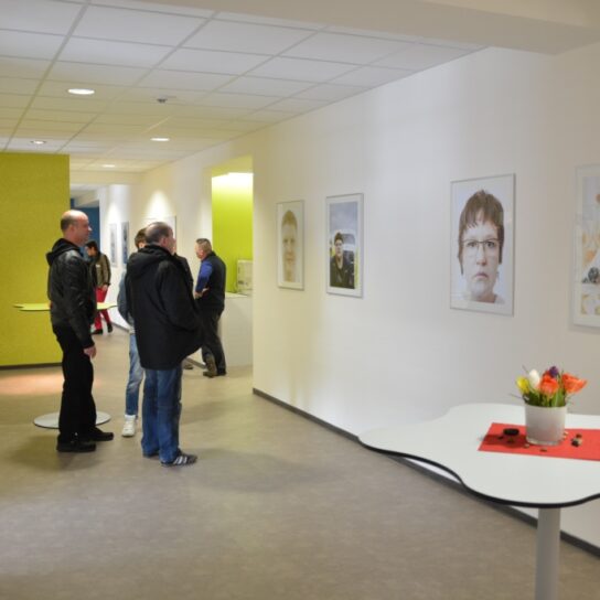 Besucher betrachten Porträtfotos in heller Galerie; Flur im Gebäude mit grünen Akzenten und Stehtisch mit Blumen.