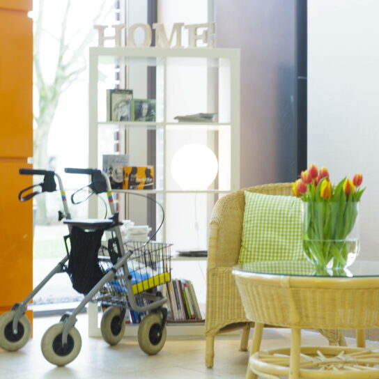 Heller, barrierefreier Wohnbereich mit Rollator, Rattanstuhl, Bücherregal und Tulpen auf Glastisch