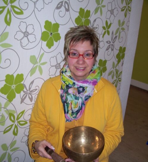 Frau mit roter Brille hält Klangschale und Klöppel in Innenraum mit Blumenmuster – Meditation und Entspannung