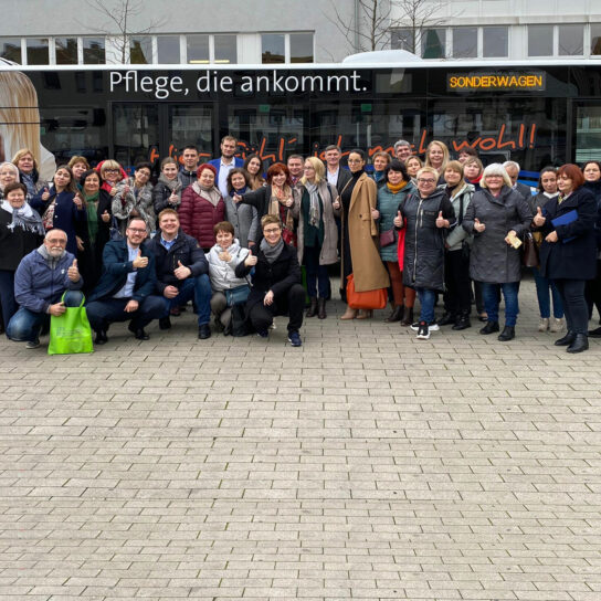 Gruppe von Pflegekräften vor Bus mit Slogan 'Pflege, die ankommt' – gemeinsames Foto mit Daumen hoch