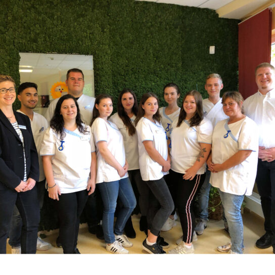 Pflegeteam in weißen Kitteln posiert vor grüner Mooswand – freundliche Pflegekräfte im Teamfoto