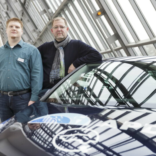 Zwei Männer neben blauem Firmenwagen im gläsernen Atrium eines modernen Gebäudes.