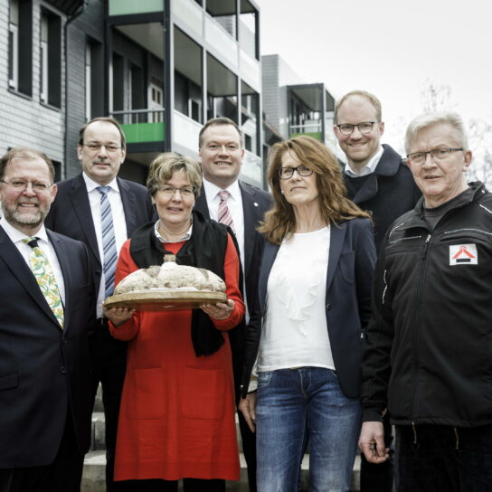 Gruppe vor neuer Wohnanlage: Frau hält Brot und Salz zur Einweihung; Verantwortliche des Bauprojekts posieren draußen.