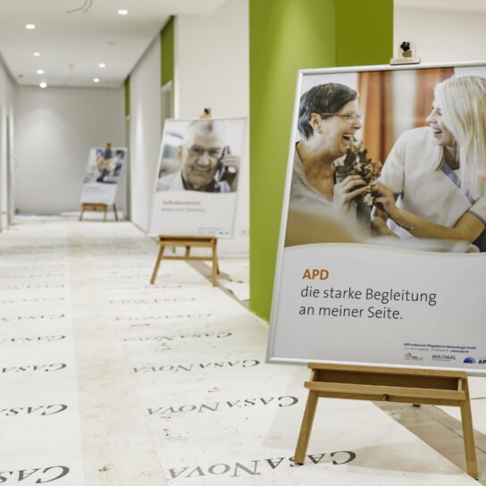 APD Pflegedienst Plakat im Flur eines Pflegezentrums – Pflegekraft und Seniorin lachen, weitere Aufsteller hinten.
