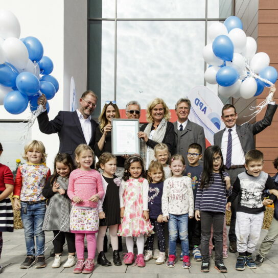 Gruppenfoto mit Kindern und Erwachsenen bei Feier vor modernem Gebäude, blaue und weiße Luftballons, Urkunde wird gezeigt.
