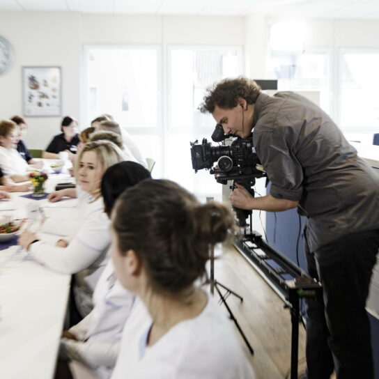 Videograf filmt Pflegekräfte bei Teammeeting im Klinikbüro, Kamera auf Slider, Besprechung am langen Tisch.