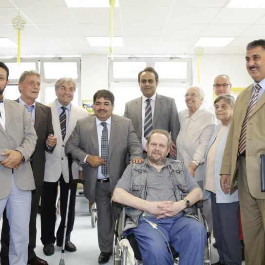 Gruppenfoto in Pflegeeinrichtung: Senioren und Gäste posieren mit Mann im Rollstuhl im Gemeinschaftsraum.