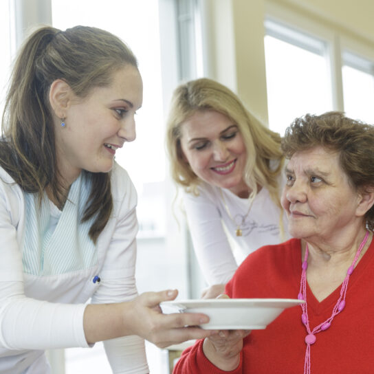 Pflegerinnen reichen Seniorin Essen im Pflegeheim – fürsorgliche Altenpflege und Betreuung beim Mittagessen.