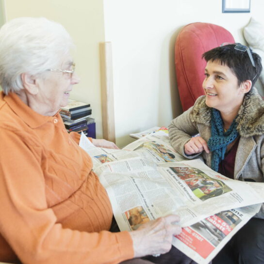 Pflegerin besucht Seniorin zu Hause, liest mit ihr Zeitung und spricht – freundliche häusliche Seniorenbetreuung.