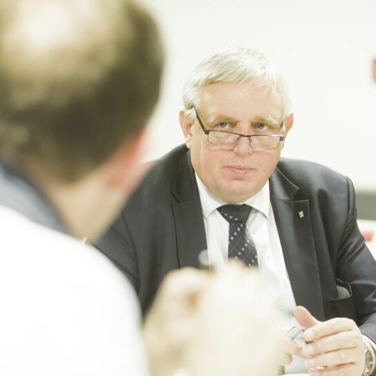 Geschäftsmann mit grauen Haaren und Brille im Anzug führt Gespräch mit Kollegen bei Meeting in heller Büroumgebung