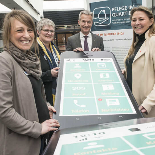 Vier Personen präsentieren digitale Pflege-App: übergroßes Smartphone bei 'Pflege im Quartier' Veranstaltung.