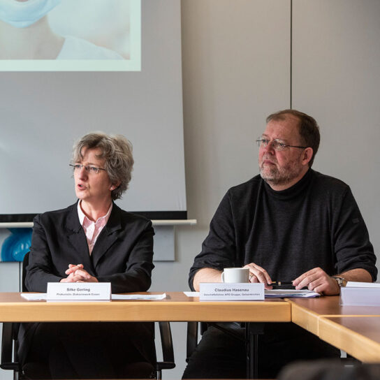 Zwei Fachleute bei einer Besprechung im Konferenzraum, vor Leinwand; Namensschilder, Notizen und Kaffeetasse.
