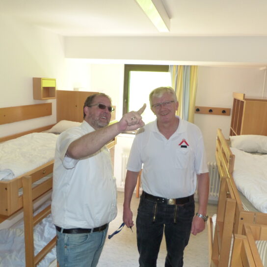 Zwei Männer besichtigen ein helles Hostel-Mehrbettzimmer mit Holz-Stockbetten und weißer Bettwäsche; einer zeigt auf Details.
