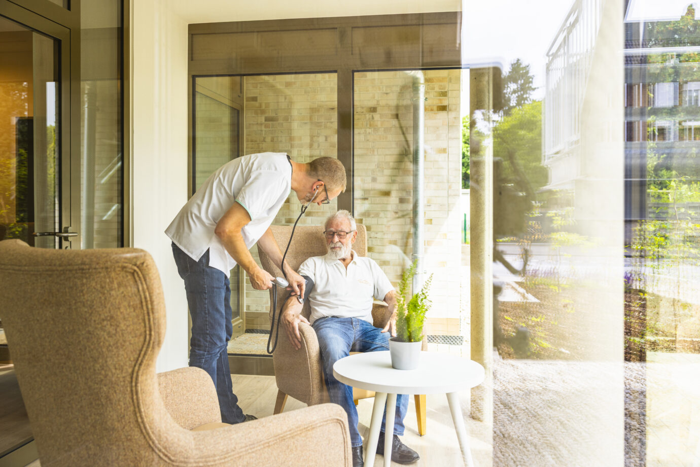 Pflegekraft misst Senior den Blutdruck im hellen Pflegeheim, fürsorgliche Seniorenbetreuung im Gemeinschaftsraum