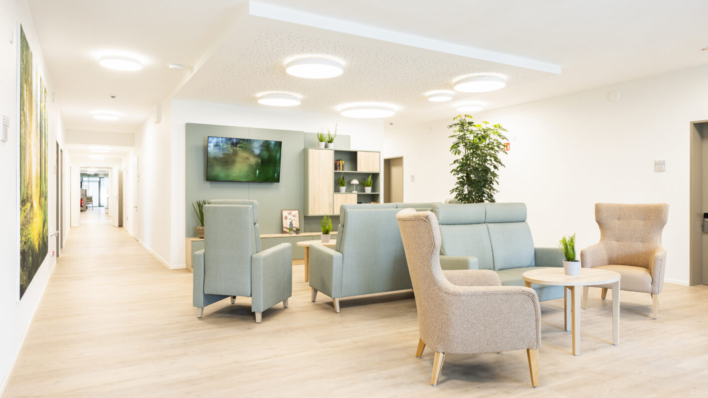 Heller, moderner Wartebereich mit bequemen Sesseln, Sofa, TV und Pflanzen in Klinik oder Pflegeheim.