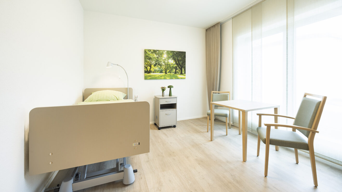 Helles, barrierefreies Pflegezimmer mit Pflegebett, Tisch und Stühlen, bodentiefes Fenster, Holzboden.
