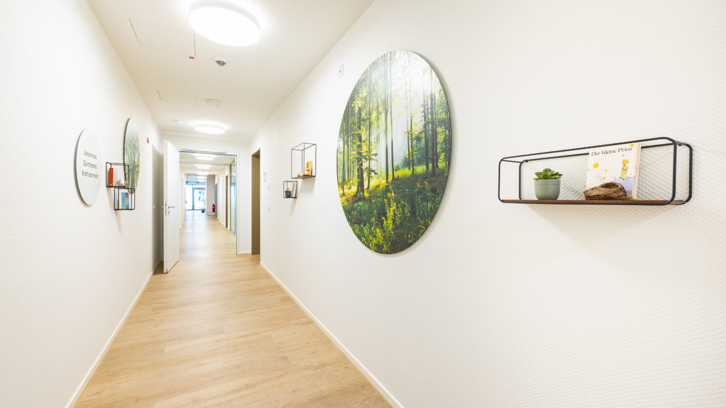 Heller, moderner Praxisflur mit Holzfußboden, rundem Waldmotiv an der Wand und Regalen mit Pflanze und Buch.