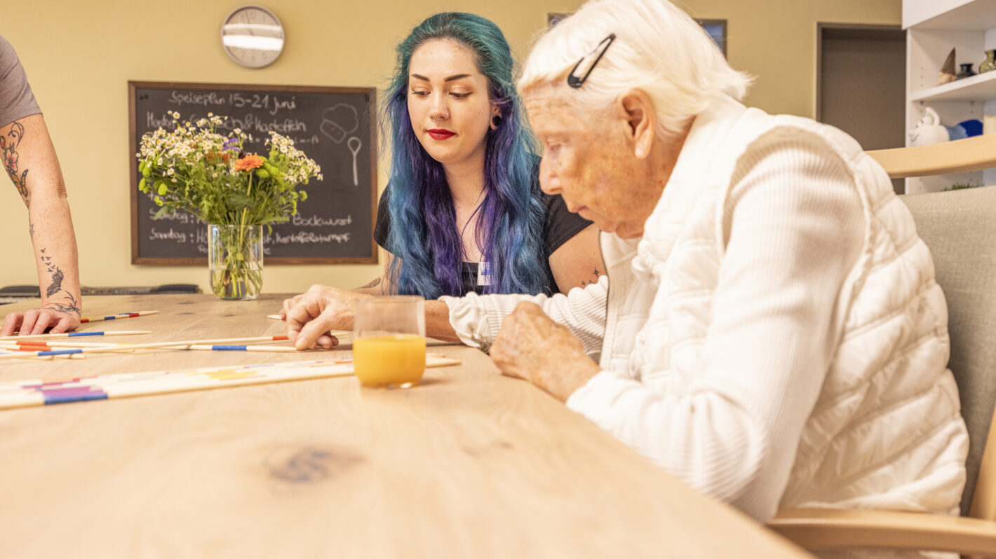 Betreuerin spielt mit Seniorin Mikado im Pflegeheim, Aktivierung und Betreuung am Tisch mit Saft und Blumen.
