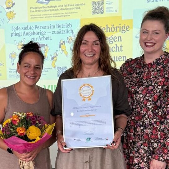Team von drei Frauen erhält Auszeichnung: Urkunde und Blumen vor Plakatwand zur Unterstützung pflegender Angehöriger.