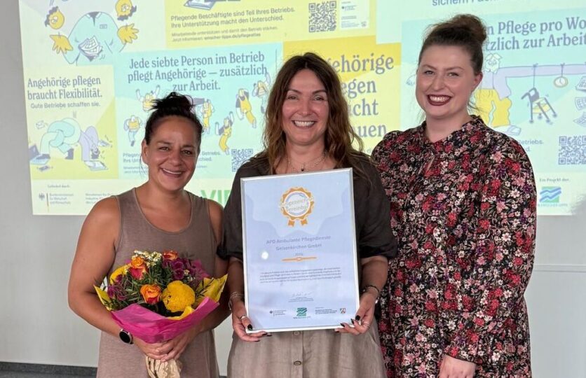 Team von drei Frauen erhält Auszeichnung: Urkunde und Blumen vor Plakatwand zur Unterstützung pflegender Angehöriger.