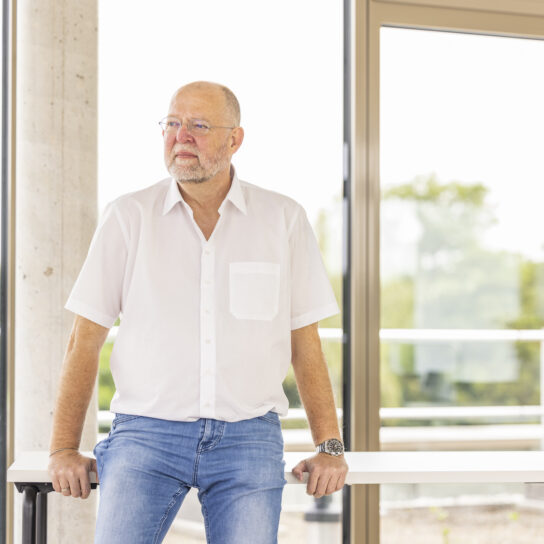Älterer Mann mit Brille und Bart im weißen Hemd lehnt an Tisch vor großen Fenstern im modernen Büro, nachdenklicher Blick.