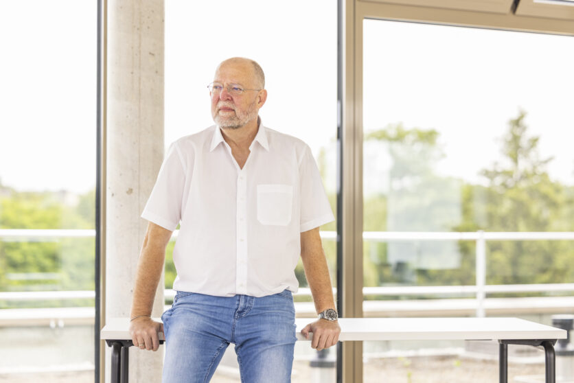 Älterer Mann mit Brille und Bart im weißen Hemd lehnt an Tisch vor großen Fenstern im modernen Büro, nachdenklicher Blick.